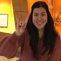 Image of Emily Prines sitting in front of skeleton display case in the UT Anthropology department, giving the Hook Em Horns sign