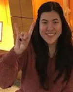 Image of Emily Prines sitting in front of skeleton display case in the UT Anthropology department, giving the Hook Em Horns sign